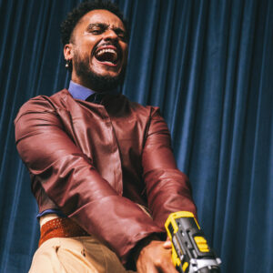  Felipe Julio, de camisa azul e jaqueta de couro. Ele segura uma furadeira amarela e parece gritar.