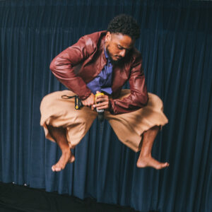 Felipe Julio, de camisa azul e jaqueta de couro. Ele olha diretamente para a câmera. Sua mão esquerda apoia a lateral da cabeça enquanto a mão direita segura uma furareira amarela em direção ao seu crânio.