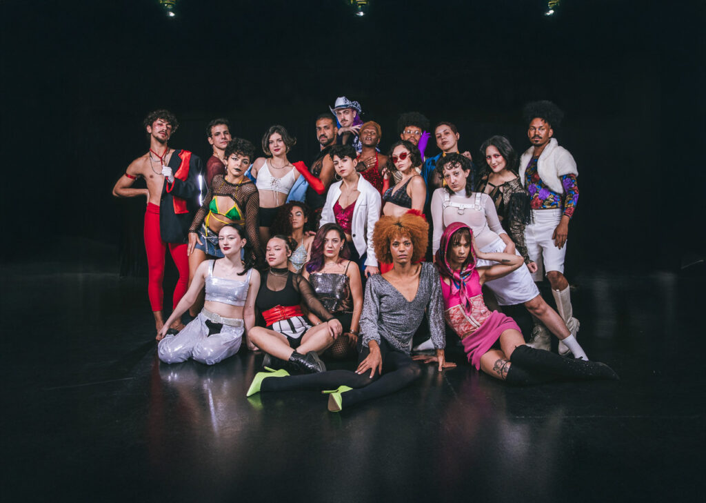 Elenco do espetáculo ZONA 21 mostra artistas LGBTQIAPN+ com corpos, etnias e gêneros diversidicades. Eles, elas, elus vestem roupas coloridas, com diferentes texturas, como rendas e peças com brilho.